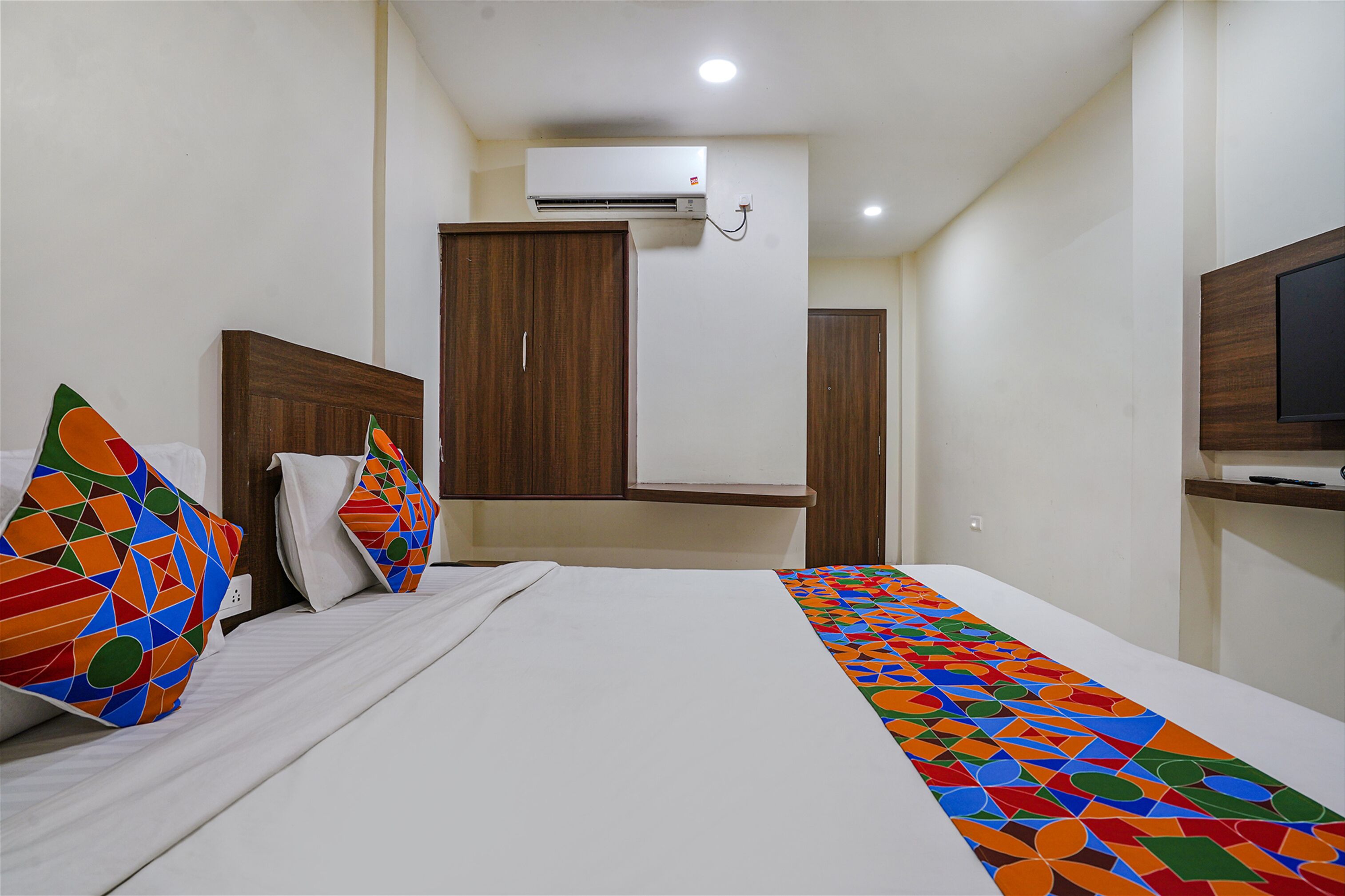 A hotel room with a bed, colorful pillows, a TV, and an air conditioning unit.