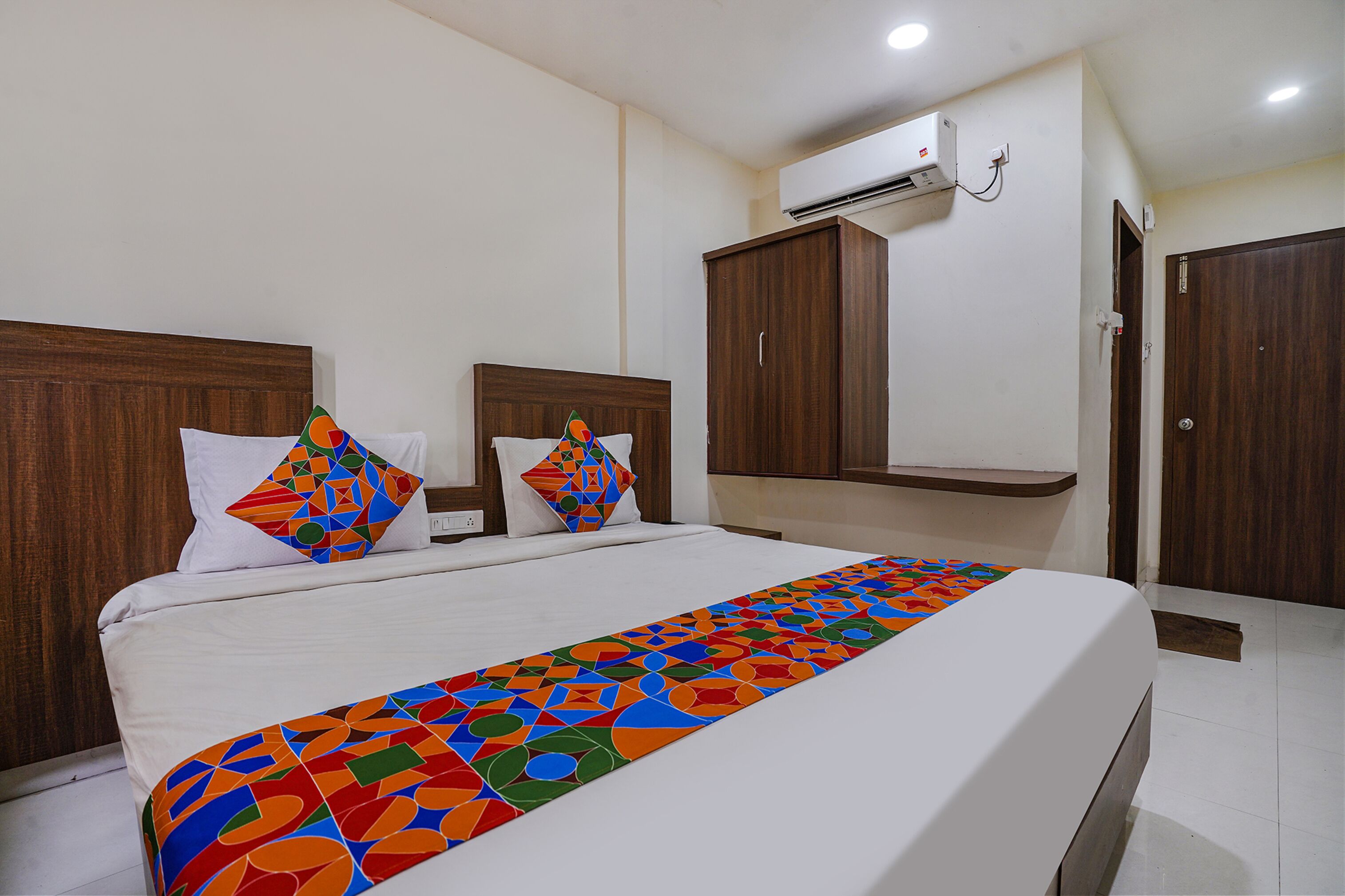 Habitación de hotel con una cama grande, dos almohadas, una colcha de colores, cabecera de madera, armario y equipo de aire acondicionado.