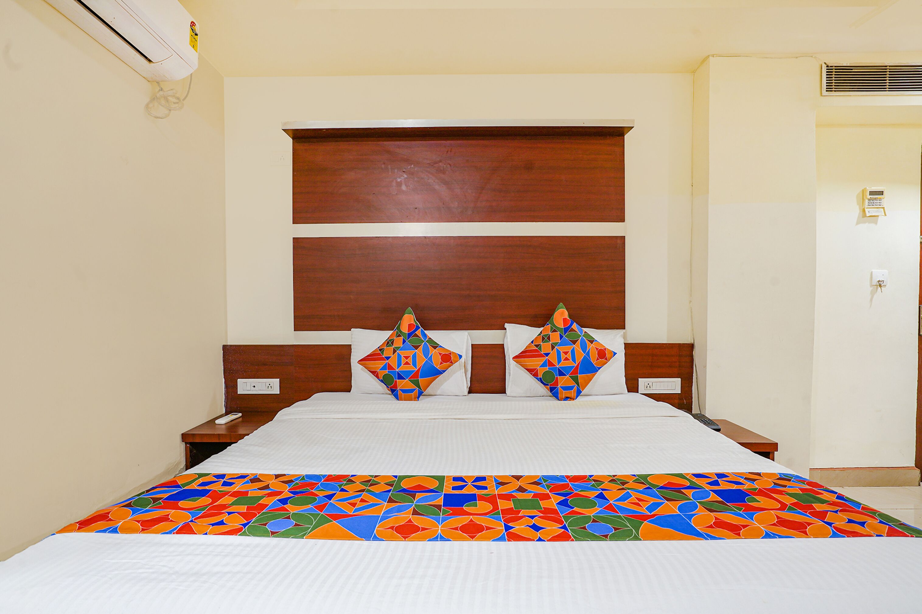 A hotel room with a large bed, wooden headboard, two decorative pillows, a bedside table, and a colorful bedspread.