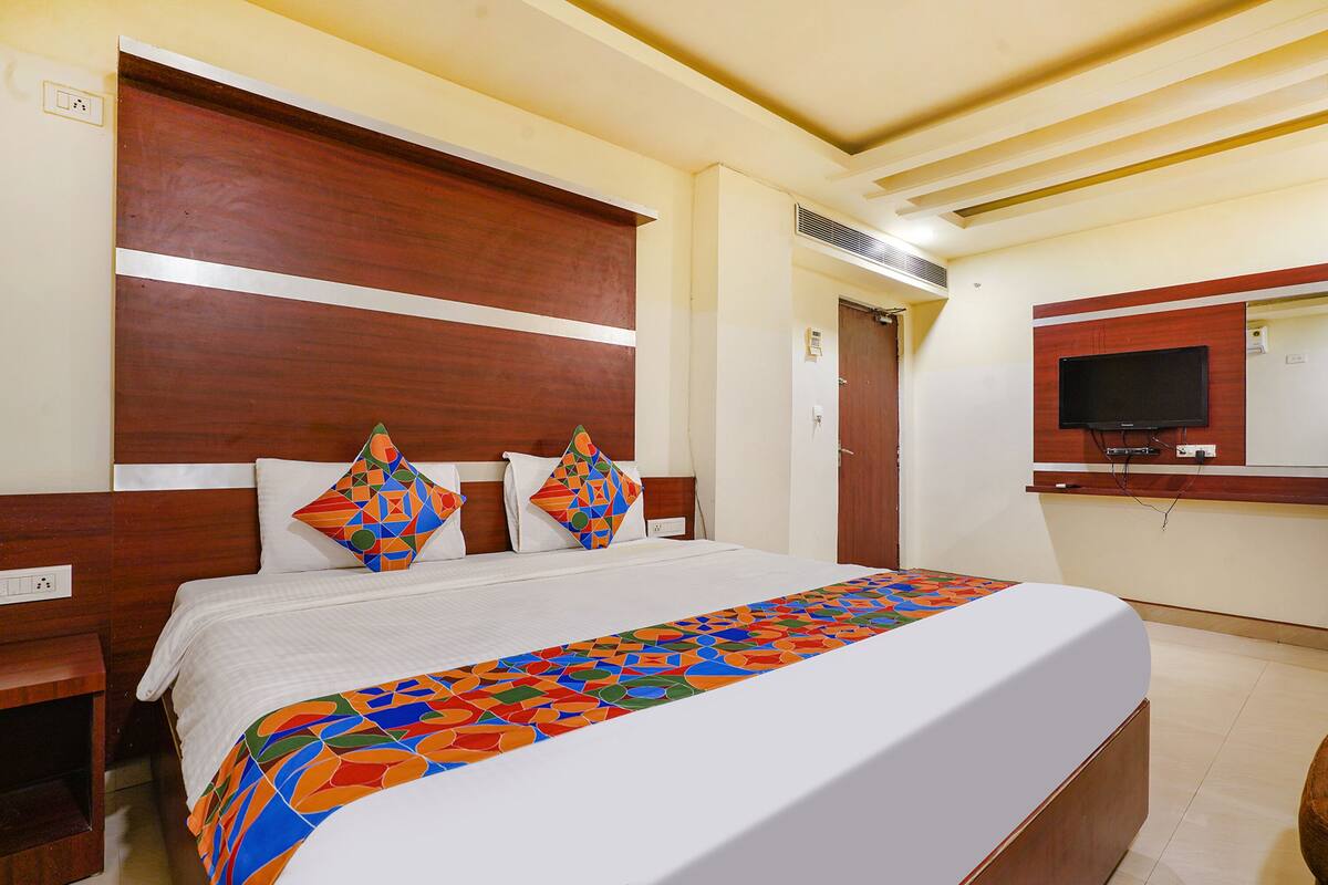 A hotel room with a large bed, wooden headboard, two decorative pillows, a colorful bedspread, a television on a wooden cabinet, and a mirror above the bed.
