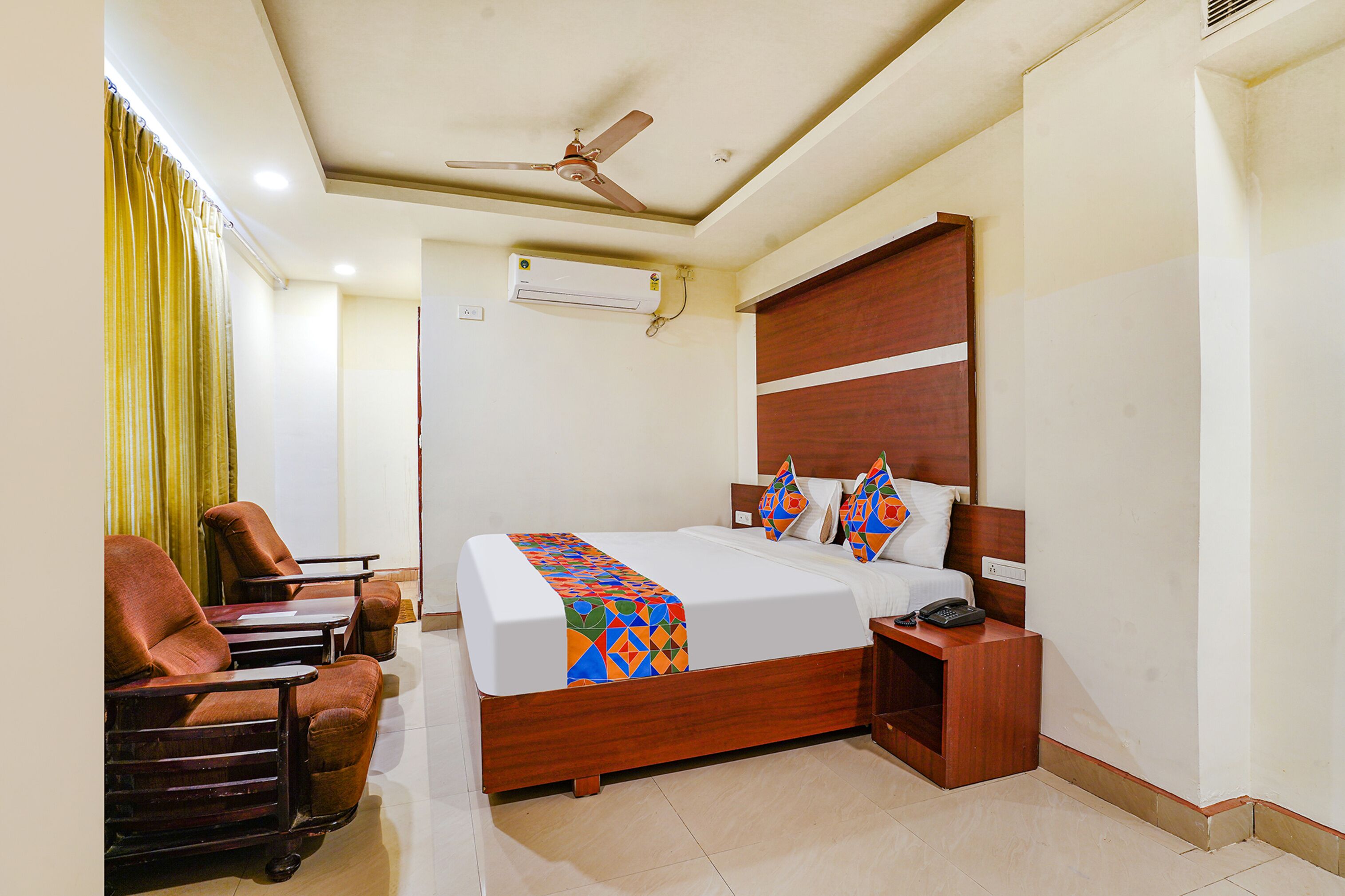 A hotel room with a bed, a desk, a chair, a nightstand, a telephone, a ceiling fan, and an air conditioning unit.