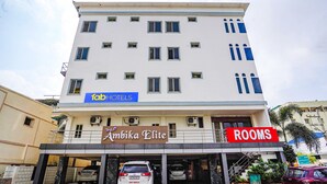 Um hotel com vários andares, com uma placa indicando 'Ambika Elite' e 'Quartos' no andar térreo. Vários carros estão estacionados em frente.