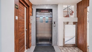 Um elevador com um aviso de 'Proibido Fumar', uma porta de madeira com placa de número e uma escada de metal com guarda-corpo.