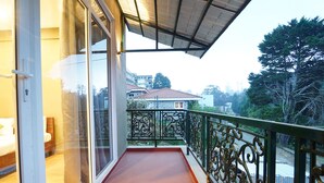 A balcony with a glass door leading to a bedroom, a metal railing, and a view of a residential area with houses and trees. - Fabhotel Hn Pear View Stays (Kodaikanal)