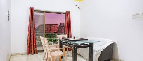 A room with a bed, a table with chairs, a window with curtains, and a view of a building outside.