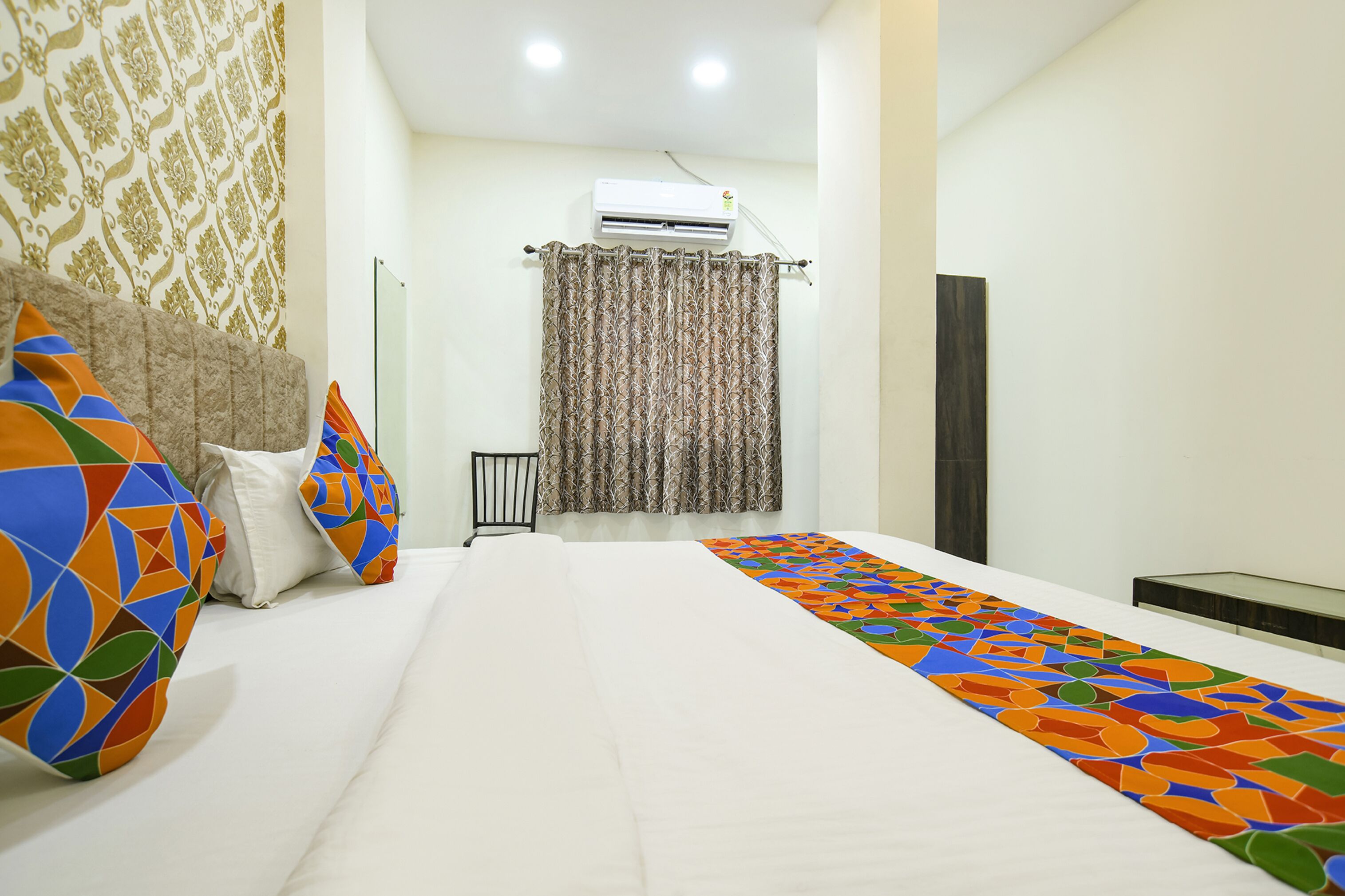 A hotel room with a bed, colorful pillows, a patterned curtain, an air conditioner, and a bedside table.