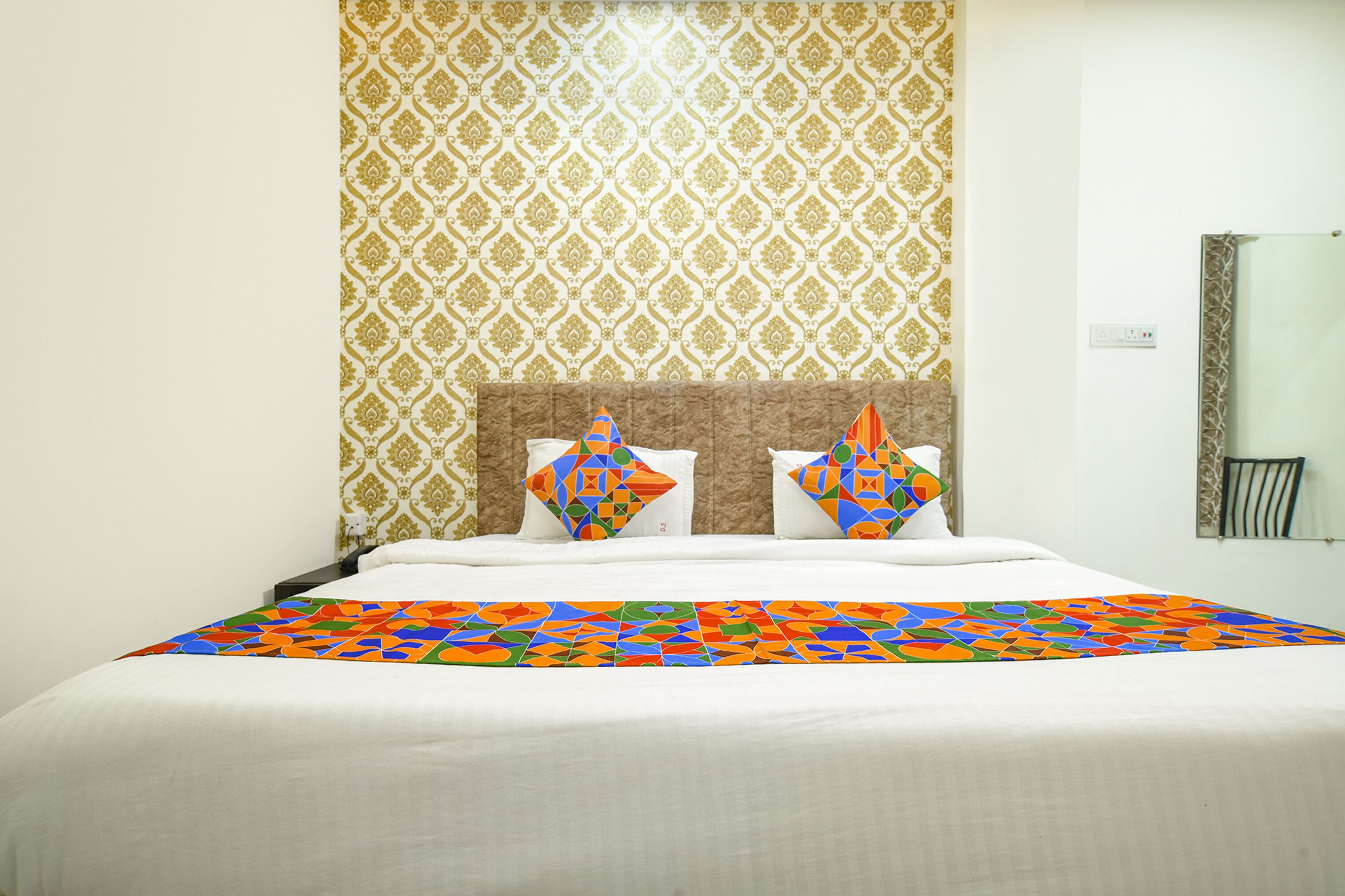 Una cama bien hecha con una colcha de colores y estampado geométrico, y dos almohadas decorativas. La habitación tiene un papel tapiz estampado y una pared blanca lisa con un espejo y una mesita.