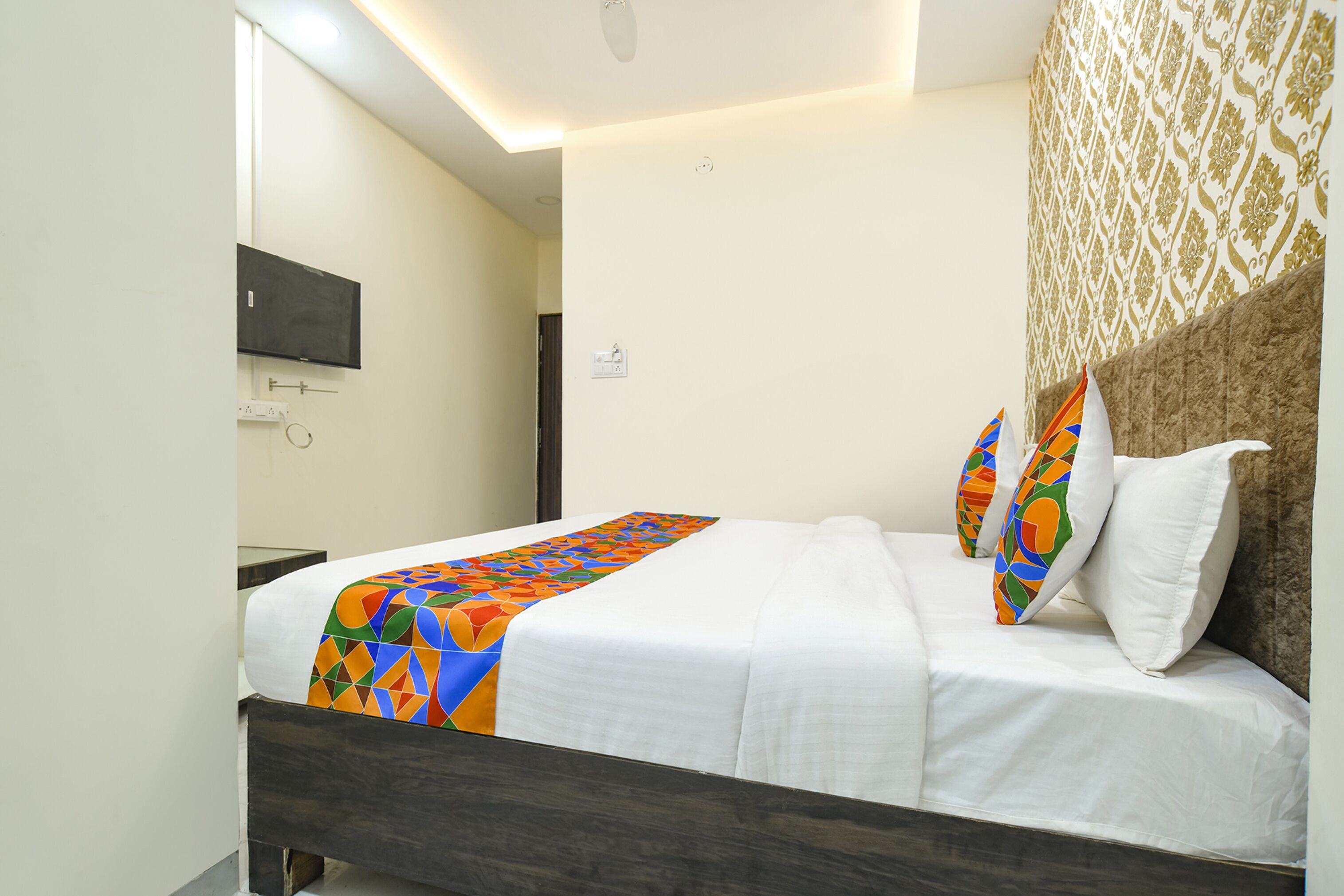 A neatly made bed with a colorful blanket and pillows, a flat-screen TV mounted on the wall, and a patterned wallpaper on the adjacent wall.