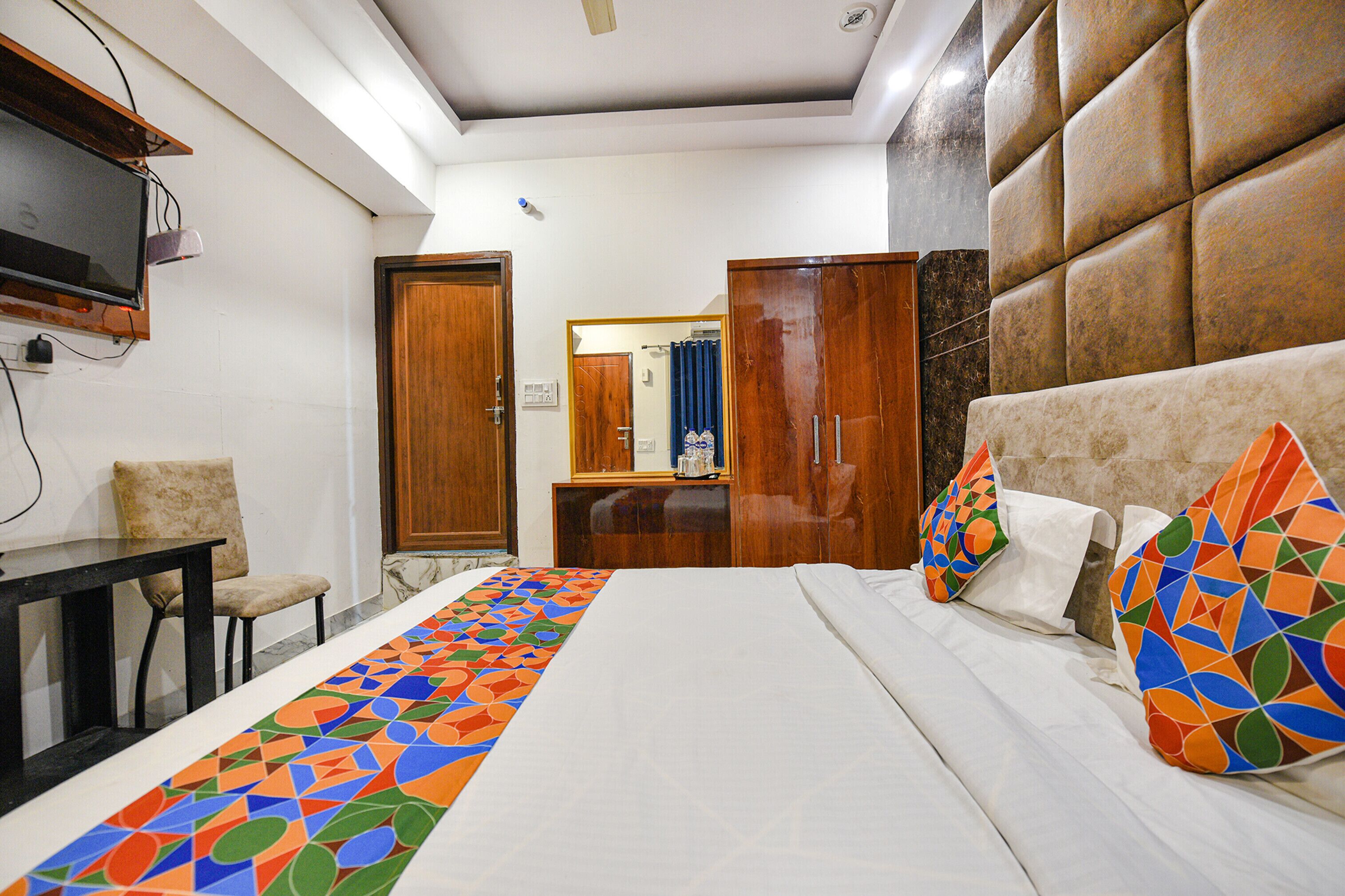 A hotel room with a bed, a TV mounted on the wall, a wooden wardrobe, and a small table with a chair.