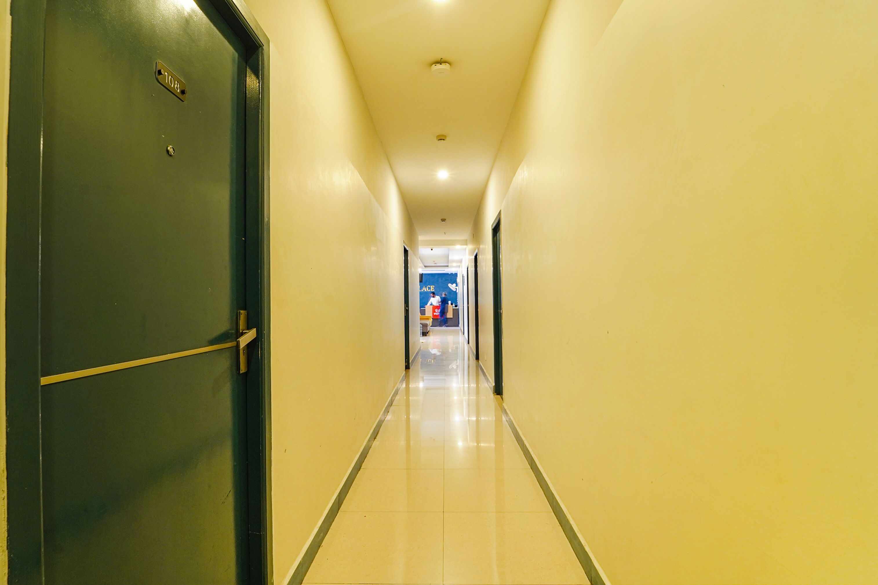 Un couloir d’hôtel avec une porte fermée sur la gauche, un sol carrelé et un panneau bleu au loin.