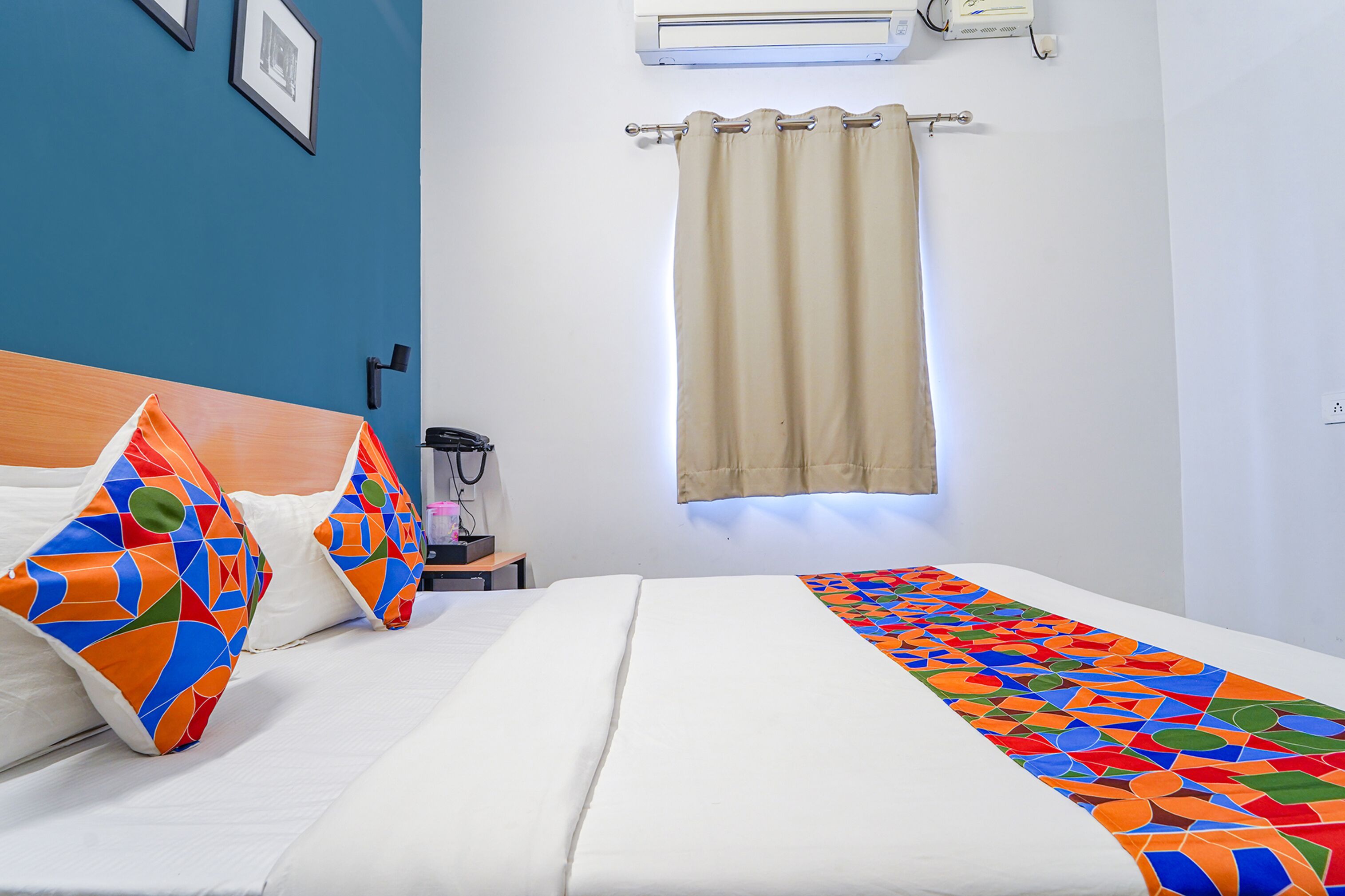 A hotel room with a bed, colorful pillows, a patterned blanket, a nightstand with a lamp, and a window with a curtain.