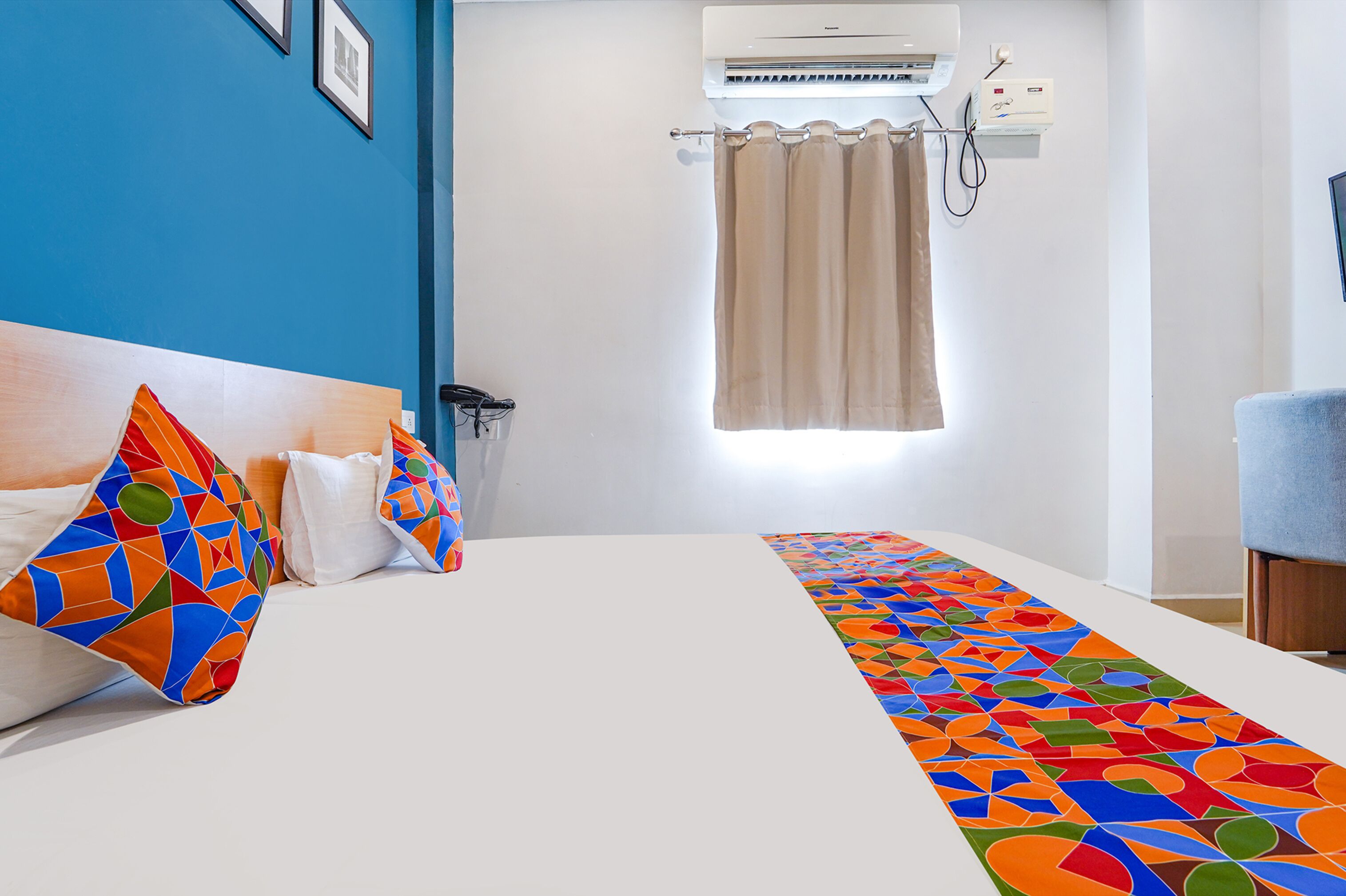 A hotel room with a bed, colorful pillows, a patterned runner, an air conditioning unit, a curtain, and a chair.