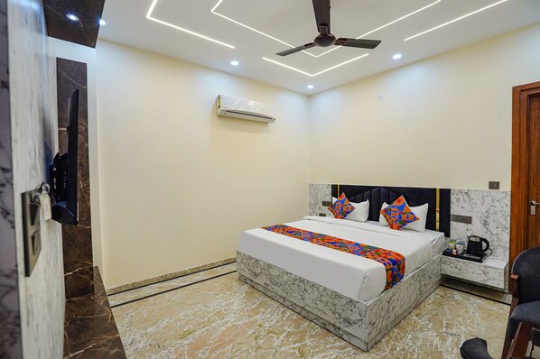 A modern hotel room with a marble floor, a bed with a white and colorful bedspread, a ceiling fan, an air conditioning unit, and a small bedside table with a kettle.