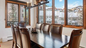 Un comedor con una mesa larga y seis sillas, ventanas amplias con vistas a edificios y árboles, y una araña con múltiples luces.