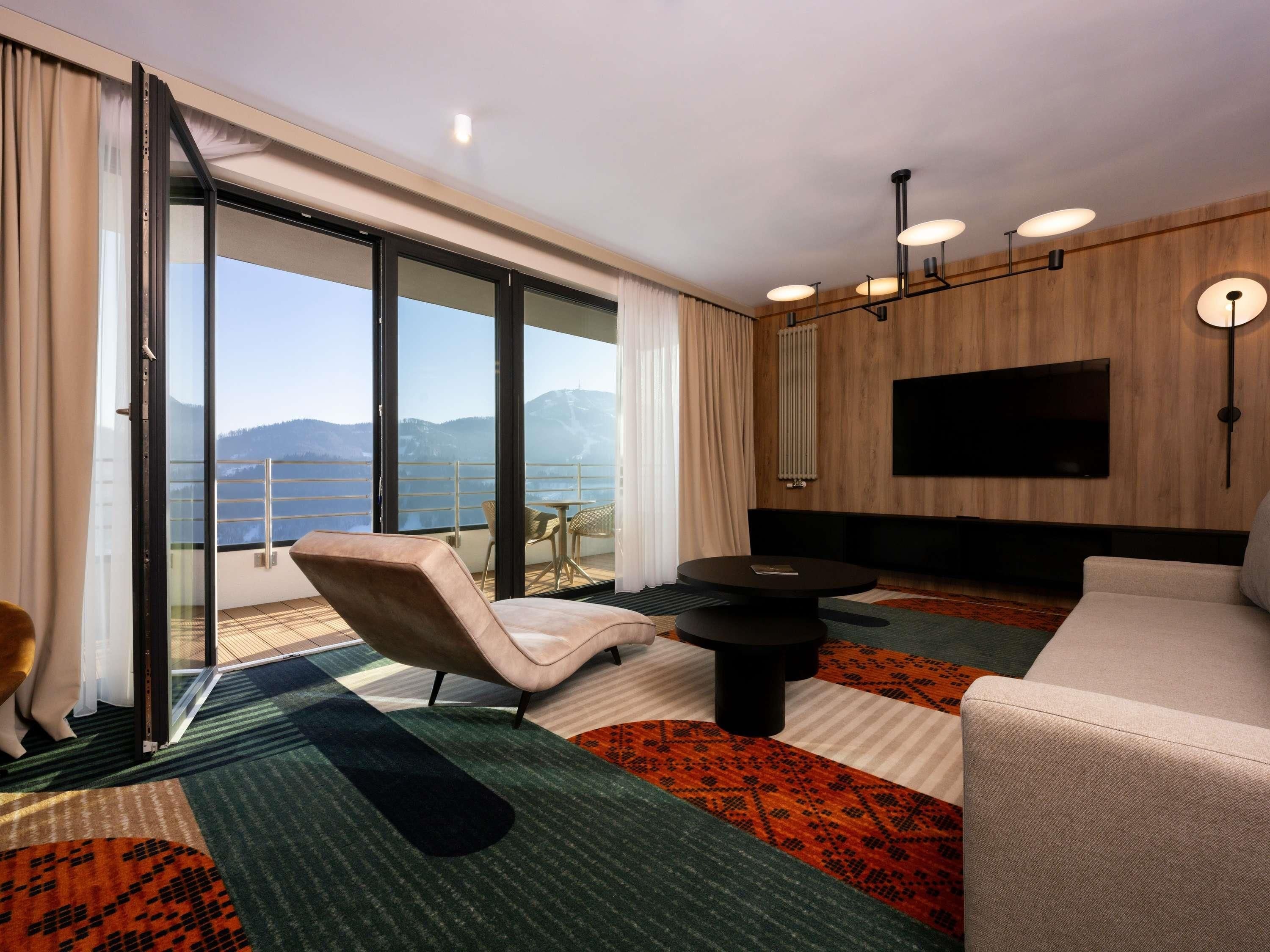 Une chambre d’hôtel moderne dotée d’une grande porte-fenêtre coulissante, d’un téléviseur à écran plat, d’un canapé, d’une table basse et d’un coin repas avec des chaises.