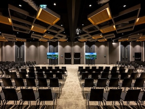 Een vergaderzaal met rijen stoelen, twee grote schermen en een modern plafondontwerp.
