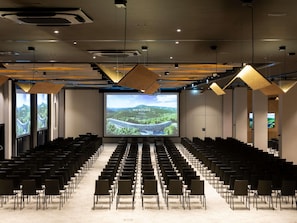 Uma sala de conferências com fileiras de cadeiras, uma tela grande exibindo um hotel e um design de teto moderno.
