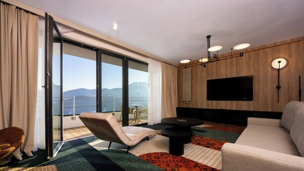Une chambre d’hôtel moderne, dotée d’un canapé, d’un fauteuil et d’une table à manger. Un grand téléviseur est fixé au mur, et la chambre offre une vue sur les montagnes.