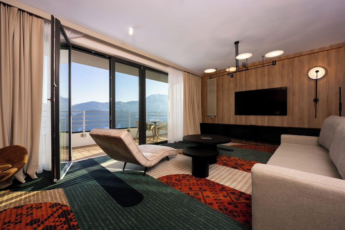 Une chambre d’hôtel moderne, dotée d’un canapé, d’un fauteuil et d’une table à manger. Un grand téléviseur est fixé au mur, et la chambre offre une vue sur les montagnes.