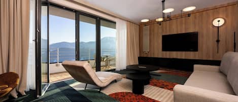 A modern hotel room with a sofa, armchair, and a dining table. A large TV is mounted on the wall, and the room has a view of the mountains.