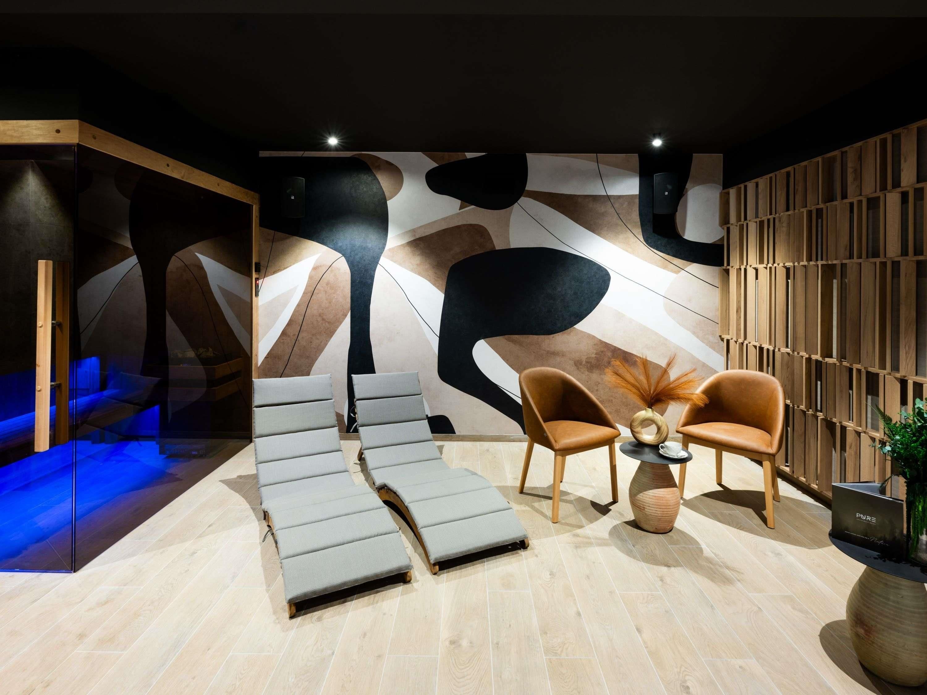 A modern spa room with a geometric wall design, a seating area with two chairs, and a massage table.