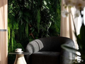 Una habitación con un sofá gris oscuro, una mesita redonda con una taza y platillo blancos, y una pared cubierta de follaje verde exuberante.