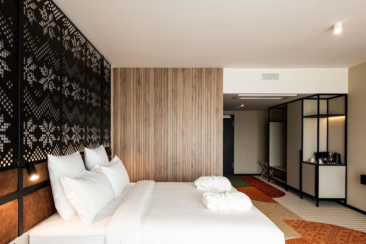 Une chambre d’hôtel moderne avec un grand lit, un mur revêtu de panneaux en bois, un panneau décoratif noir et un petit coin salon.