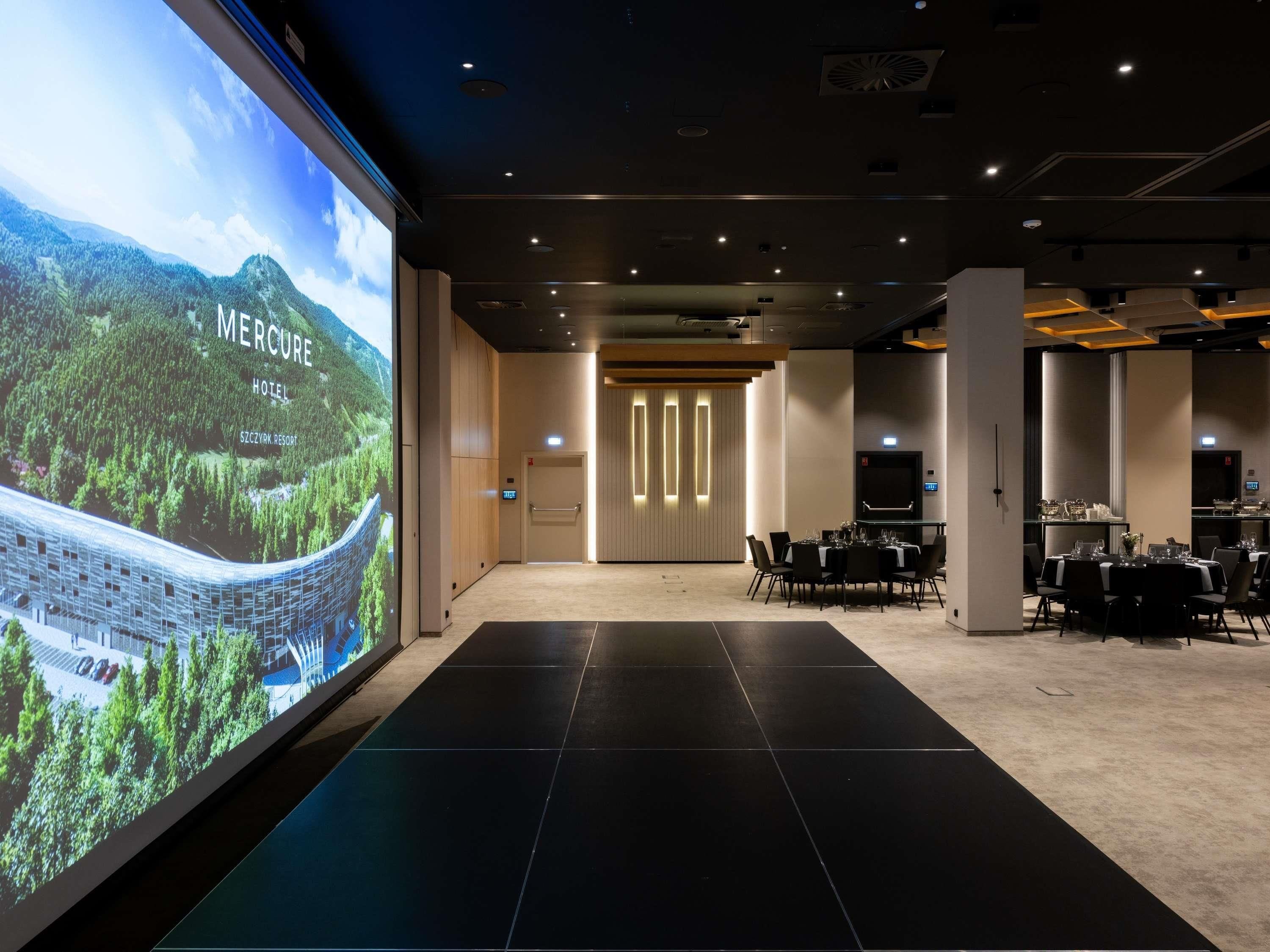 Un hall d’hôtel doté d’un grand écran diffusant des images de paysages, d’un comptoir d’accueil et d’un espace repas avec des tables préparées pour les clients.