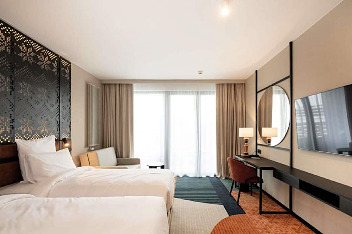 Une chambre d’hôtel avec deux lits, un bureau, une chaise, un grand miroir et une vue sur l’extérieur par la fenêtre.