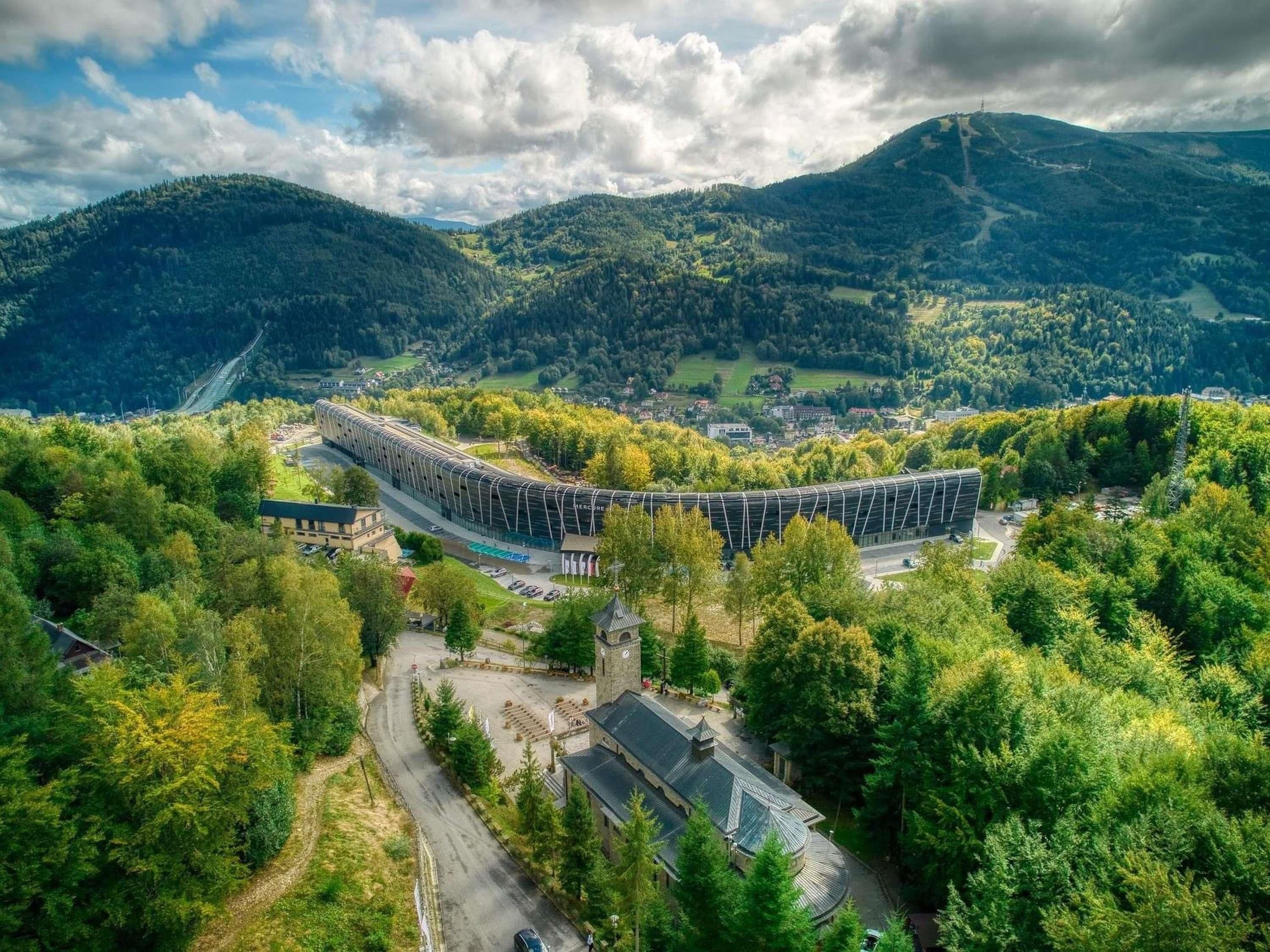 A modern building with a curved roof, surrounded by lush greenery and hills.