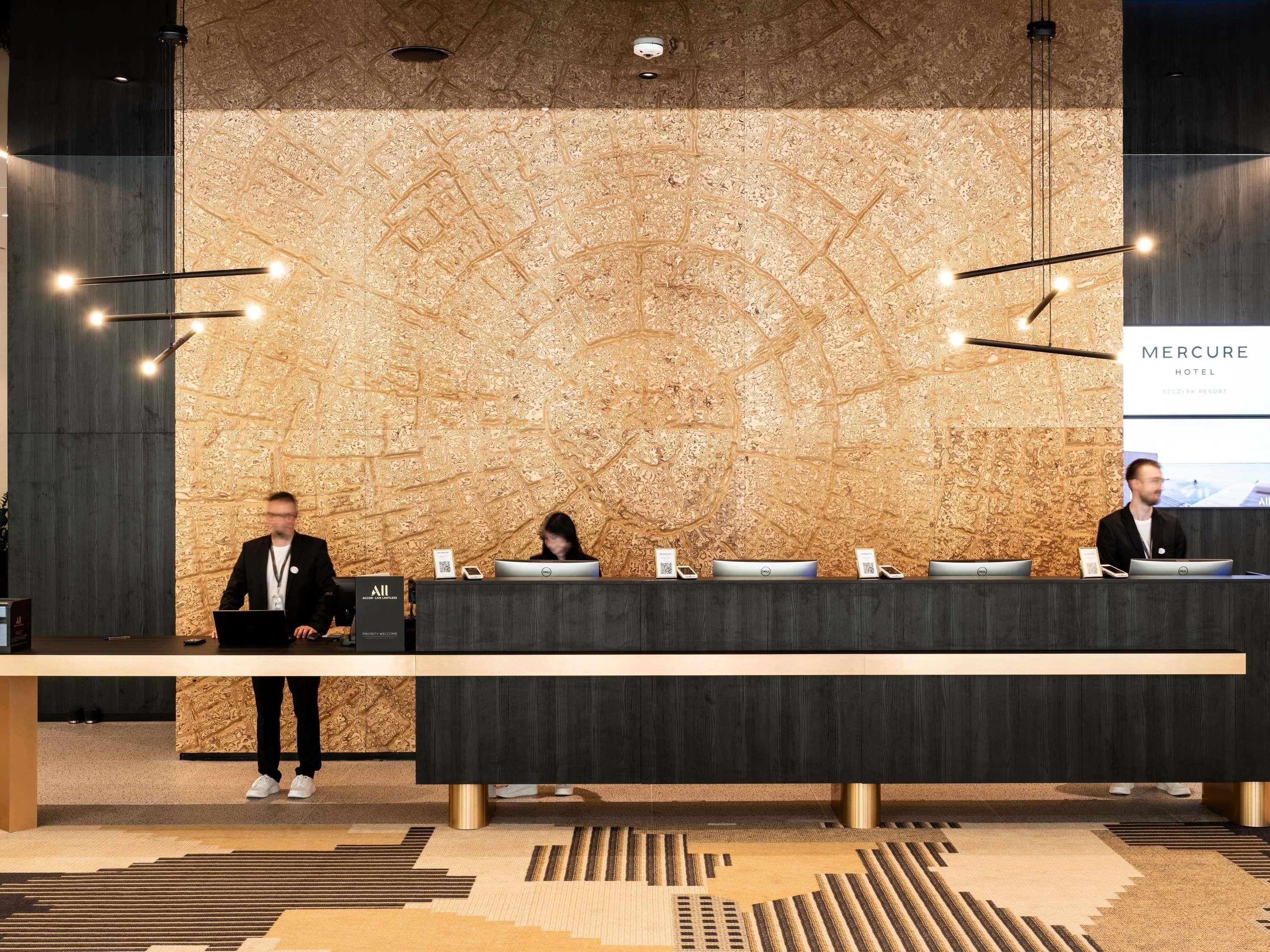 A reception desk with a wooden wall backdrop, a digital screen displaying 'Mercure Hotel,' and a modern chandelier.