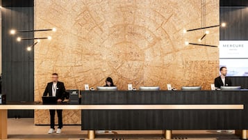A reception desk with a wooden wall backdrop, a digital screen displaying 'Mercure Hotel,' and a modern chandelier.
