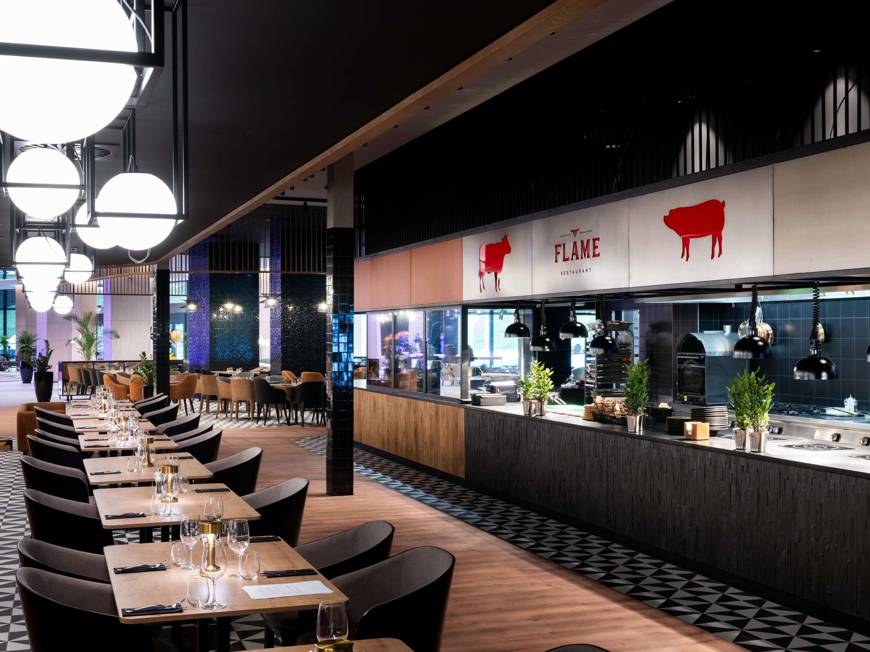 Un interior de restaurante con mesas dispuestas para comer, una zona de cocina y un diseño moderno con lámparas colgantes.