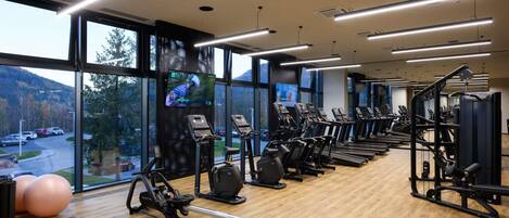 Un gimnasio con máquinas de cardio, zonas de pesas y amplias ventanas que dan a un estacionamiento y árboles.