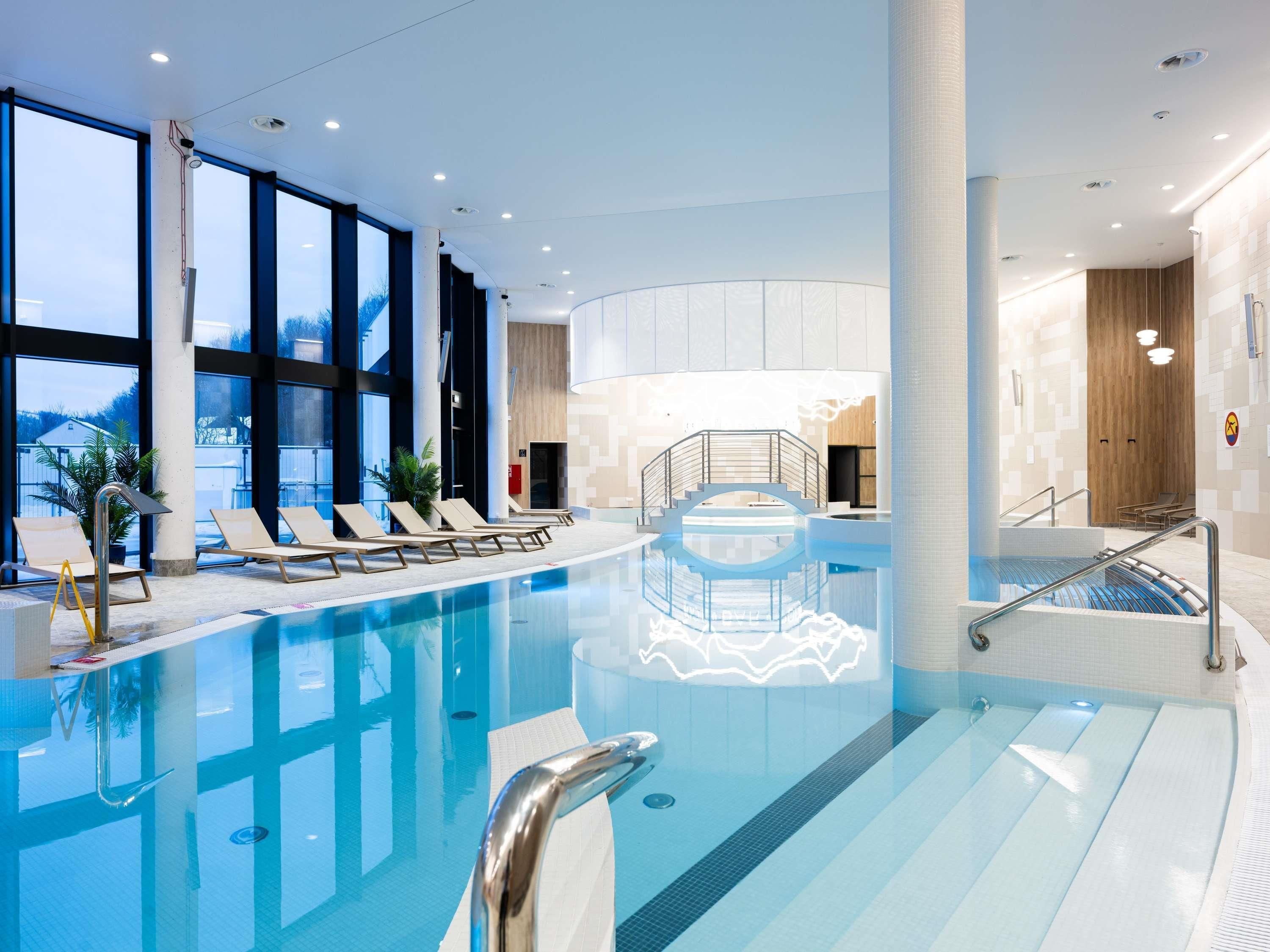 A modern indoor swimming pool with clear water, sun loungers, and a view of the outdoors.