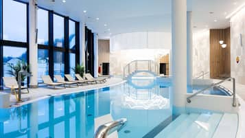 A modern indoor swimming pool with clear water, sun loungers, and a view of the outdoors.