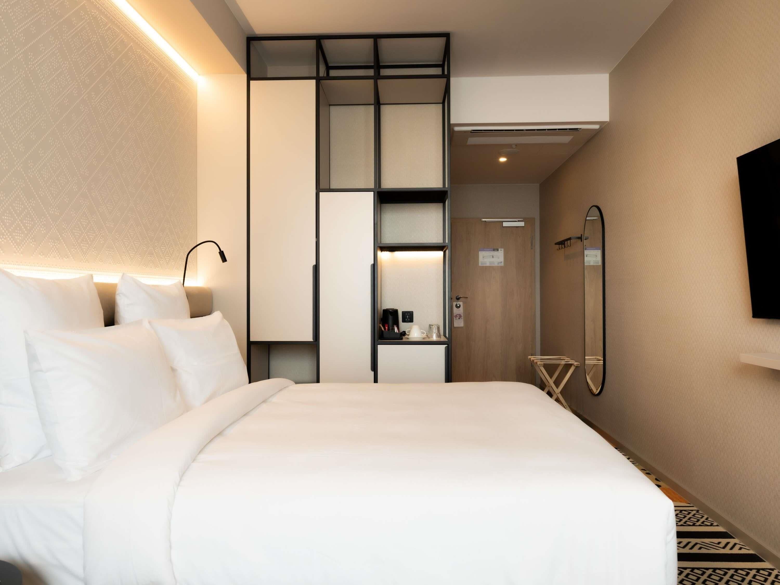 Une chambre d’hôtel équipée d’un lit, d’une table de chevet, d’une lampe, d’une armoire, d’un miroir et d’un téléviseur fixé au mur.