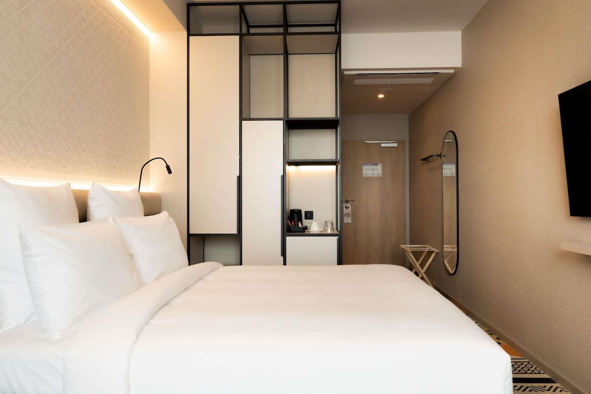 Ein Hotelzimmer mit Bett, Nachttisch, Lampe, Kleiderschrank, Spiegel und einem Fernseher an der Wand.