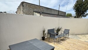 A wooden deck with a dark wicker table and chairs, surrounded by a low wall and a stone building in the background. - Chez Fanf et Nanie (Isigny-sur-Mer)