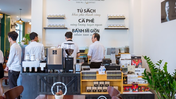 A coffee shop interior with a counter, coffee machines, and shelves with coffee products. - BIG HOTEL (Hue)