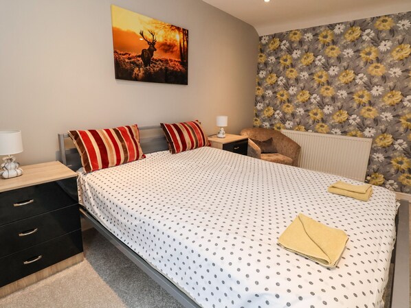 Un dormitorio con una cama, dos almohadas, una mesita de noche, una silla, un radiador y un papel tapiz con motivos florales.