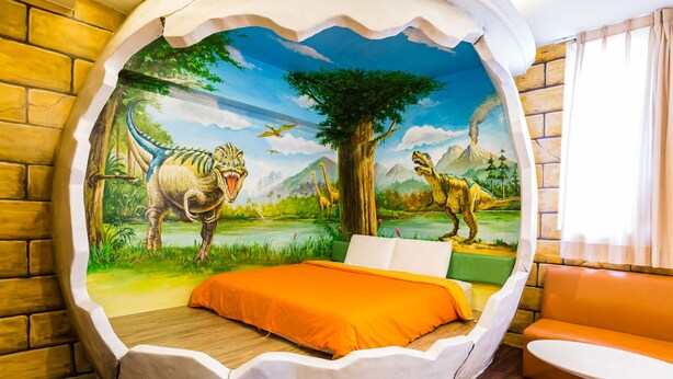 Een kamer met een muurschildering van dinosaurussen en een boom, een bed met een oranje sprei en een witte stoel.