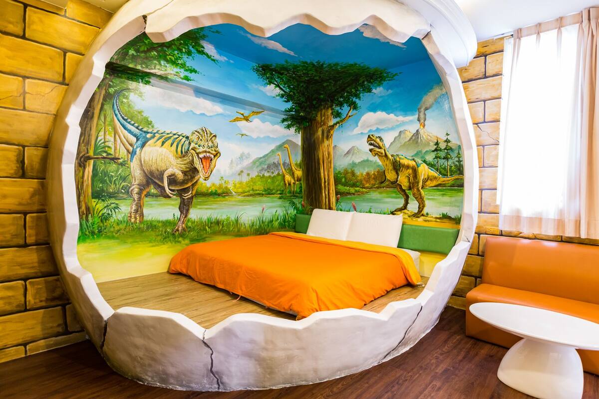 A room with a mural of dinosaurs and a tree, a bed with an orange bedspread, and a white chair.