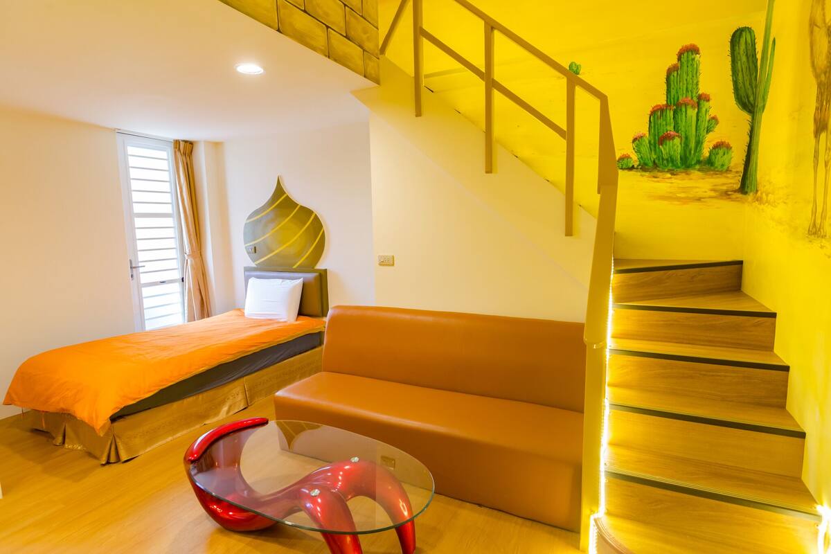 A bedroom with a bed, a sofa, a glass table, and a staircase with a yellow wall and a cactus mural.
