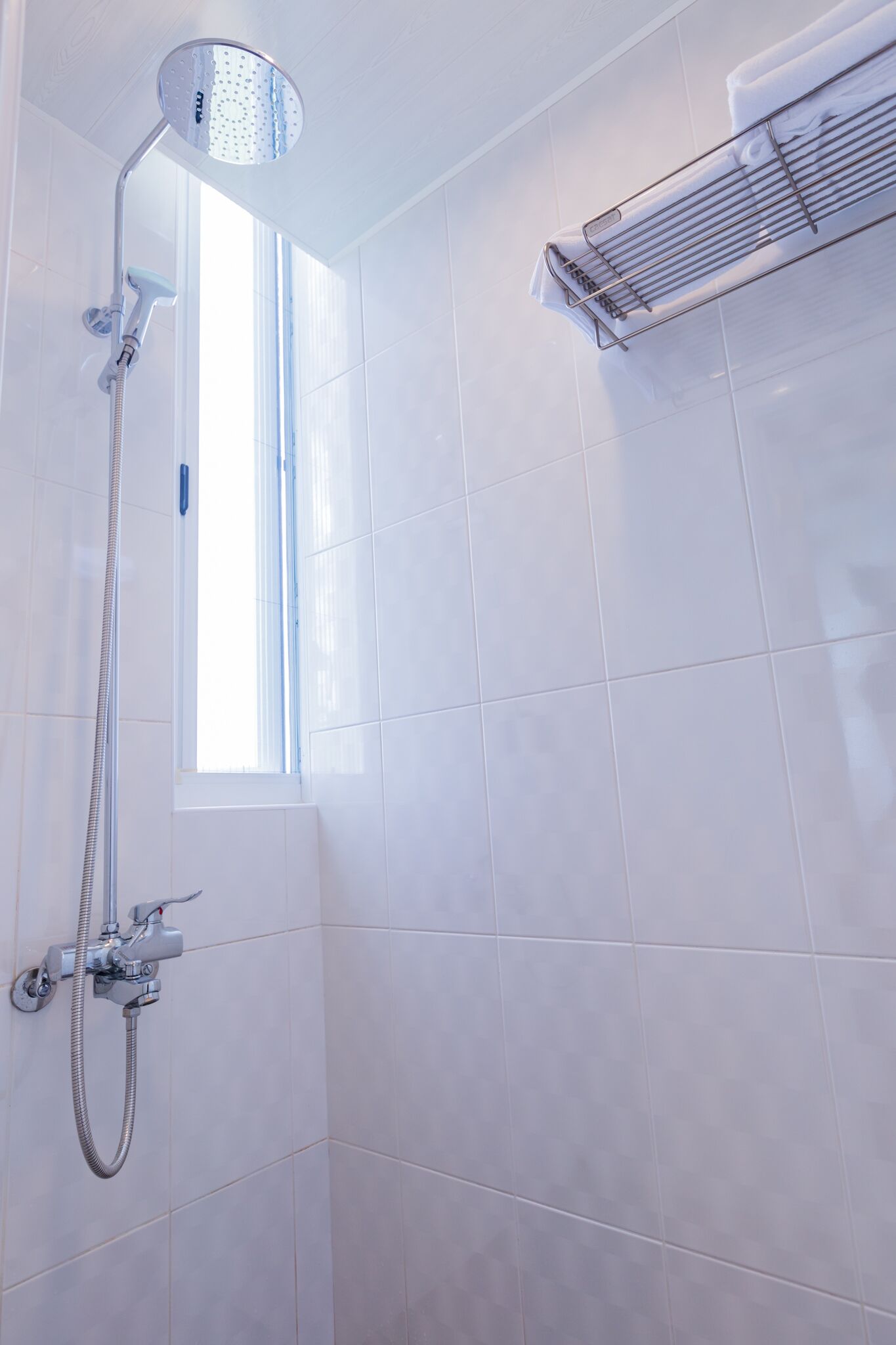 A shower with a handheld showerhead and a wall-mounted control panel, a window with blinds, and a towel rack on the wall.