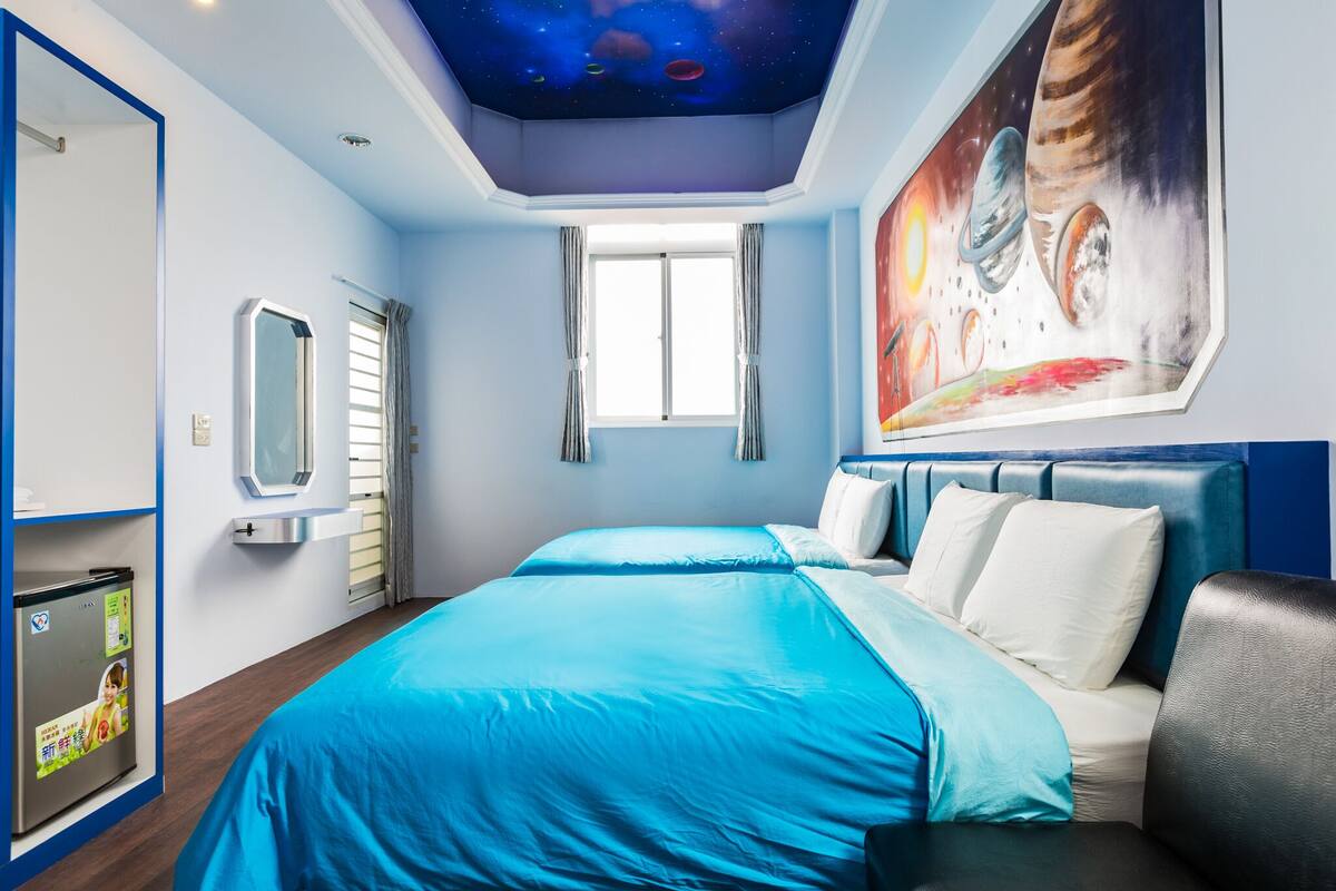 A bedroom with a bed, a window, a mirror, a refrigerator, and a wall with a space-themed mural.