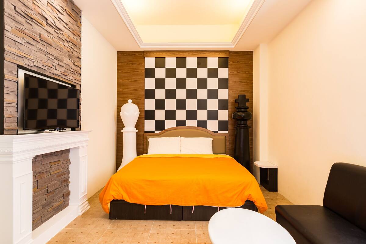 A bedroom with a checkered headboard, a flat-screen TV mounted above a fireplace, and a bed with an orange bedspread.