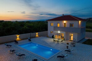 A modern two-story house with a swimming pool, outdoor seating, and a view of the surrounding landscape.