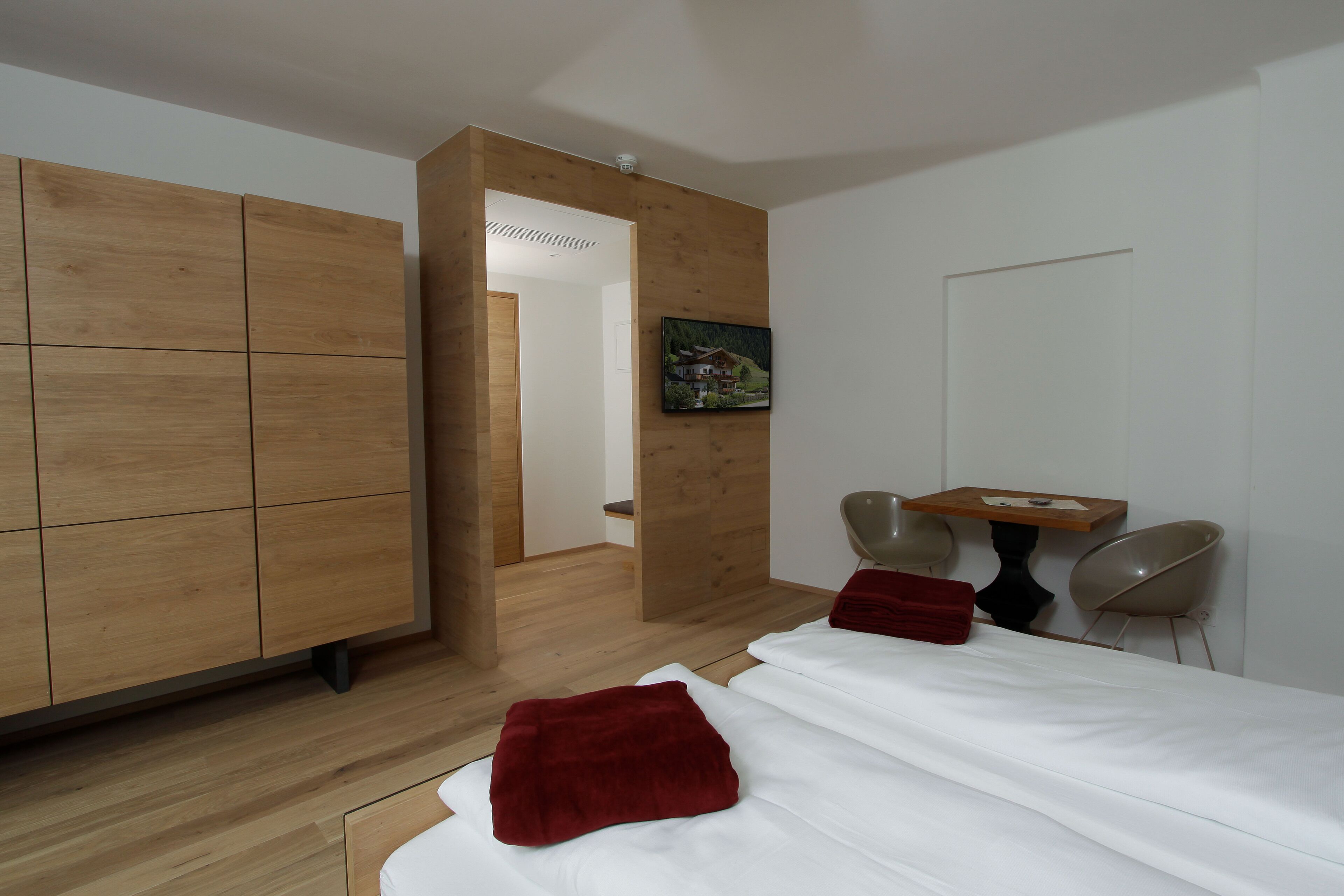 A modern bedroom with a wooden wardrobe, a bed with white bedding and red pillows, a small table, and two chairs.