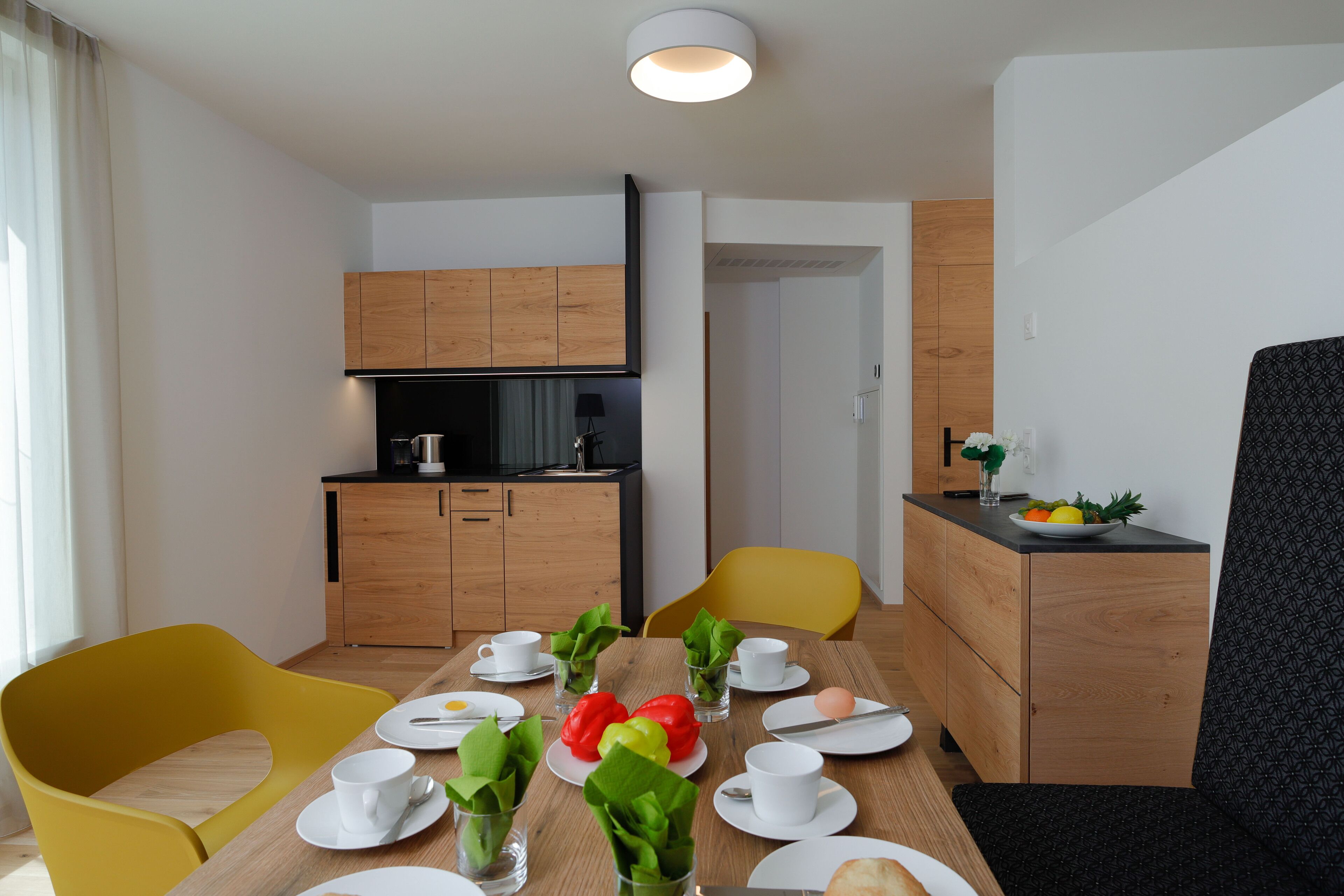 Un comedor moderno con una mesa de madera para dos personas, sillas amarillas y una pequeña cocina al fondo.
